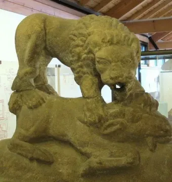 The Corbridge Lion sandstone sculpture showing a lion standing over another animal, displayed indoors beside other stone objects.
