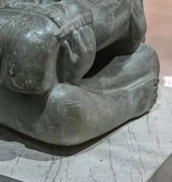 Lower body of the Las Limas Figurine seated on a marble base, with bent legs, visible feet, and scratch marks on the stone surface.