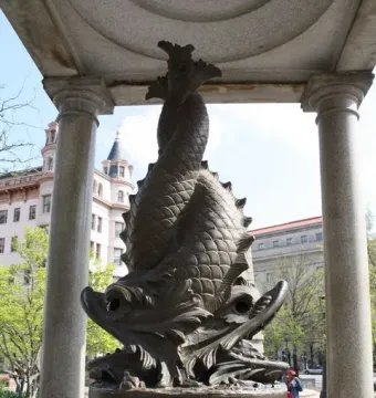 Bronze fish sculpture on the Temperance Fountain, framed by two stone columns with buildings in the background.