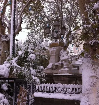 Ain El Fouara Fountain covered in snow, with sculpted figures, a street lamp on top, and snow-laden trees and fence nearby.