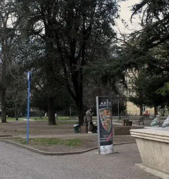 Stone fountain with metal frog statues beside a park path, with trees and a building in the background.