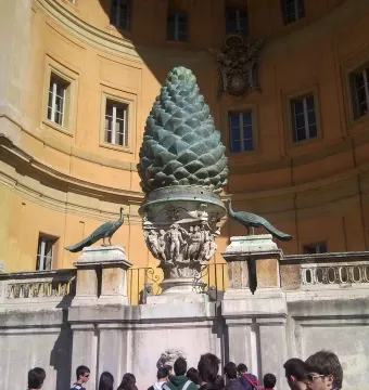 La Pigna bronze pine cone sculpture on a carved pedestal with peacock statues in front of a curved building.