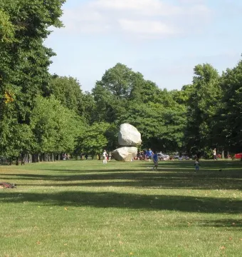 Rock on Top of Another Rock stands in a wide grassy lawn bordered by trees, with a few people visible in the distance.