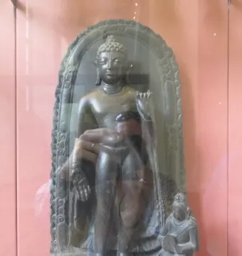 Sultanganj Buddha displayed in a glass case, with a standing figure inside an arched stele and a kneeling figure at the base.