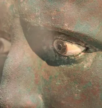 Close-up of the Meroë Head showing the eye, nose, and cheek of a weathered bronze statue with green and reddish-brown patina.