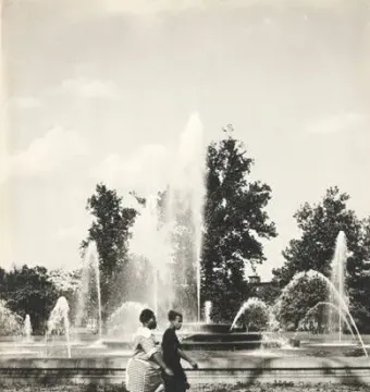 Two women walk past the Swann Memorial Fountain with water jets spraying into the air.