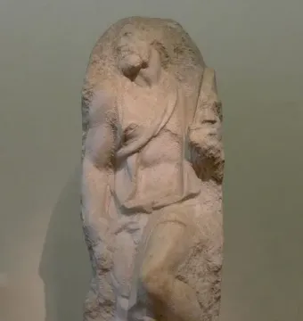 Unfinished marble sculpture of Saint Matthew emerging from stone, with a bearded figure looking upward.