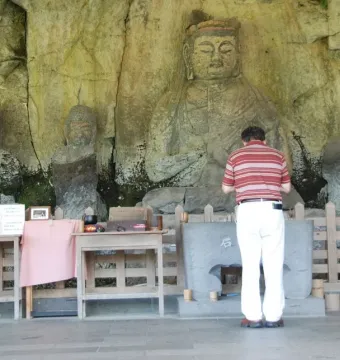 A man bows before the Usuki Stone Buddhas carved into a rock wall, with offerings placed on tables in front of the figures.