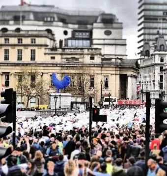 The blue Hahn/Cock sculpture stands on a pedestal in Trafalgar Square with a large crowd nearby.