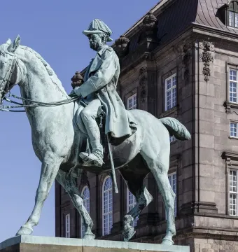 Christian IX equestrian statue in bronze against a blue sky, with a building facade behind it.