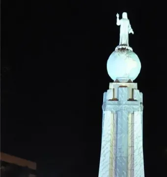 Divino Salvador del Mundo monument illuminated at night, with a robed statue standing on a glowing blue globe above a tall stone pedestal.