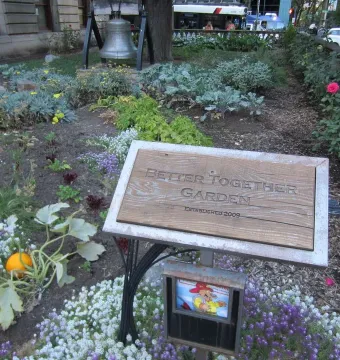 The Liberty Bell mounted in a garden bed beside a stone building and flowering plants.