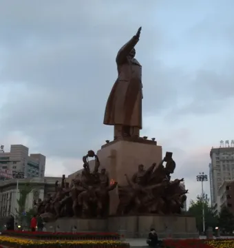 The Long Live the Victory of Mao Zedong Thought statue in a city square, with Mao raising an arm on a tall pedestal and smaller bronze figures around the base.