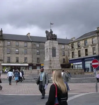 Paisley War Memorial on a tall stone pedestal in a paved town square with surrounding buildings and pedestrians.