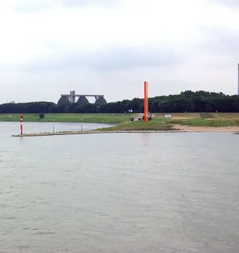 Rhine Orange sculpture standing on a grassy riverbank beside a wide river, with trees and industrial structures in the background.