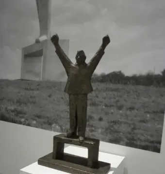 Small bronze model of the Monument to Stjepan Filipović with raised clenched fists on a pedestal, shown before a large black-and-white background photo of the monument.