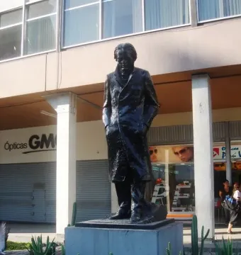 Monument to César Vallejo showing a dark metal figure in a long coat on a concrete pedestal, with cacti and buildings behind it.