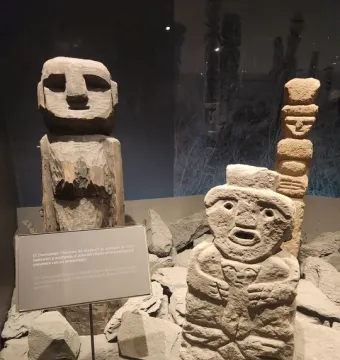 Chemamull wooden figure displayed among grey stones with smaller stone sculptures nearby.