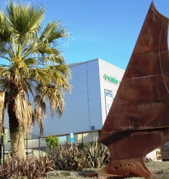 Dorna rust-colored metal sculpture with a sail-like curved shape in a landscaped area with palm trees and a white building behind it.