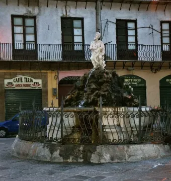 The Fountain of the Genius of Fieravecchia stands in a cobblestone square, with a weathered marble base, small statue, and surrounding metal fence.