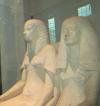 Horemheb and Amenia seated side by side in a light-colored ancient Egyptian sculpture behind glass.