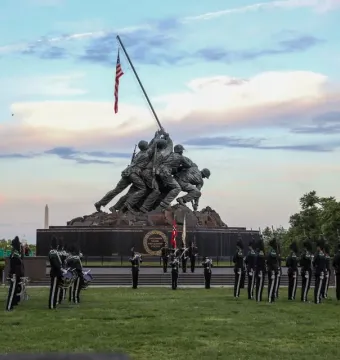 The Marine Corps War Memorial at twilight, with a military band or honor guard assembled on the lawn in front of the statue.