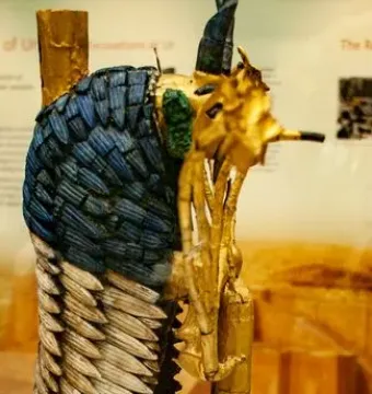 Ram in a Thicket sculpture with dark curved horns, blue and white body inlay, and a gold-colored support in a museum display.