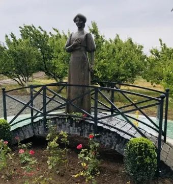 Bronze statue of Zabel Yessayan behind a black metal fence and stone bridge, surrounded by rose bushes and greenery.