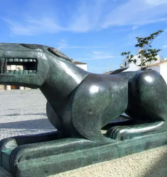 The Leona de Baena sculpture in green metal, shown in profile on a stone base in a town square.