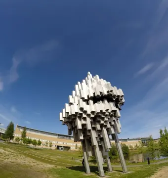 Norra skenet sculpture with clustered silver blocks beside a grassy lawn, with buildings, water, and trees in the background.