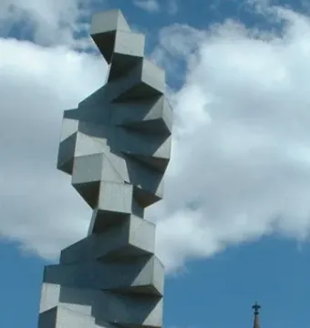 Endlose Treppe, a tall twisting gray sculpture against a blue sky, with a church spire and tree in the background.