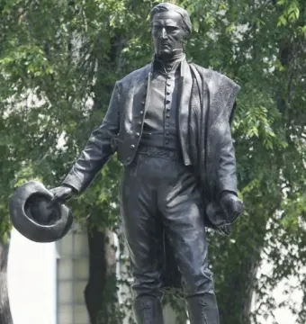 Bronze statue of General José Gervasio Artigas standing in historical clothing and holding a hat, with trees and a building behind it.