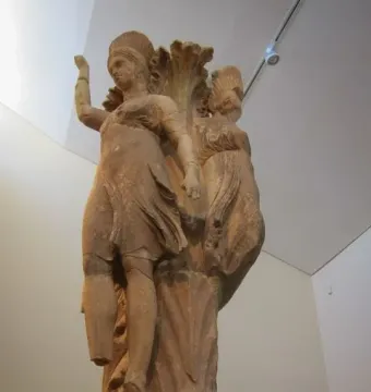 Dancers of Delphi sculpture showing two draped female figures with raised arms around a central pillar on an ornate base.