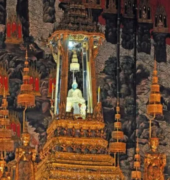 Emerald Buddha statue in a pale golden robe on an ornate golden altar, surrounded by gilded decorations and mural-covered walls.