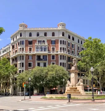 The Fountain of Hercules stands on a tiered stone base in a Barcelona square with trees and a large building behind it.