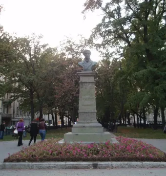 Bust of Pushkin on a tall stone pedestal in a park with pink flowers, trees, and people walking nearby.