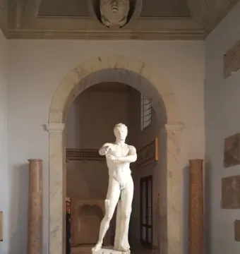 Apoxyomenos, a white marble nude male statue, stands on a pedestal in a museum room with an arched entryway and tiled floor.