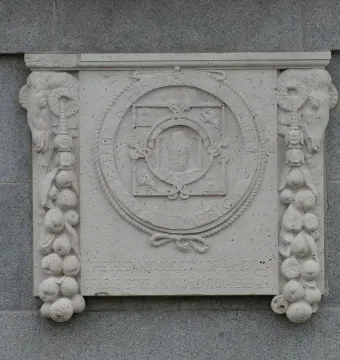 Decorative stone plaque for El Cid mounted on a granite wall, with carved ram heads, fruit clusters, and a circular crest with text.