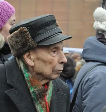 An elderly person in a fur-lined cap and striped scarf stands near the Solovetsky Stone in snowy weather.