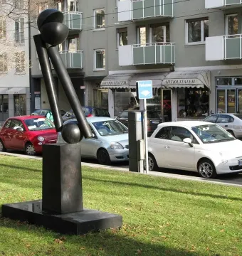 Große Zwei V, a dark abstract sculpture with cylindrical and spherical forms on a rectangular base in a grassy urban park.