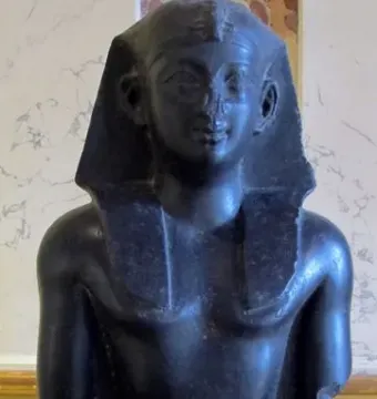 Upper torso of a Ptolemaic king statue in dark stone, with a nemes headdress and missing arms.