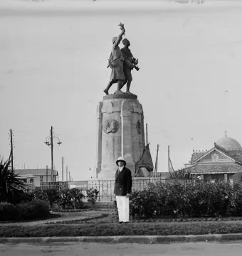 The Demba et Dupont monument stands on a stone plinth outdoors, with a man in the foreground and buildings behind it.