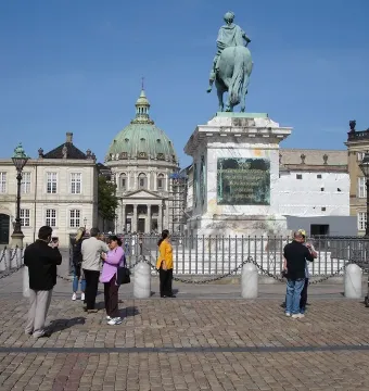 Frederick V equestrian statue on a tall marble pedestal in a sunny square, with people nearby and a green-domed church in the background.