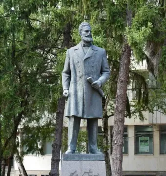 Monument to Hristo Botev: a bronze-colored statue of a bearded man holding a rolled document on a tall stone pedestal among trees.