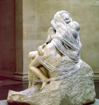 Marble sculpture of two figures embracing, draped with thin strings over a textured stone base.