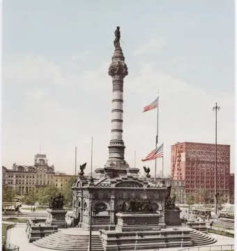 Soldiers\' and Sailors\' Monument rising from a stepped base in a landscaped park, with American flags nearby and city buildings in the background.
