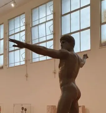 Artemision Bronze, a nude bronze male statue posed with one arm extended and the other drawn back in a museum display.