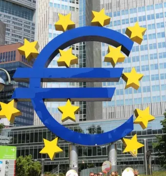 Blue euro sculpture with yellow stars in front of modern glass buildings, with people gathered below it.