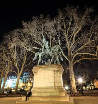 Illuminated bronze monument of Charlemagne and His Leudes with a rider and two standing figures on a tall stone pedestal at night.