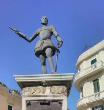 Bronze statue of John of Austria in historical armor on a tall stone pedestal, with one arm extended and a sword in the other hand.